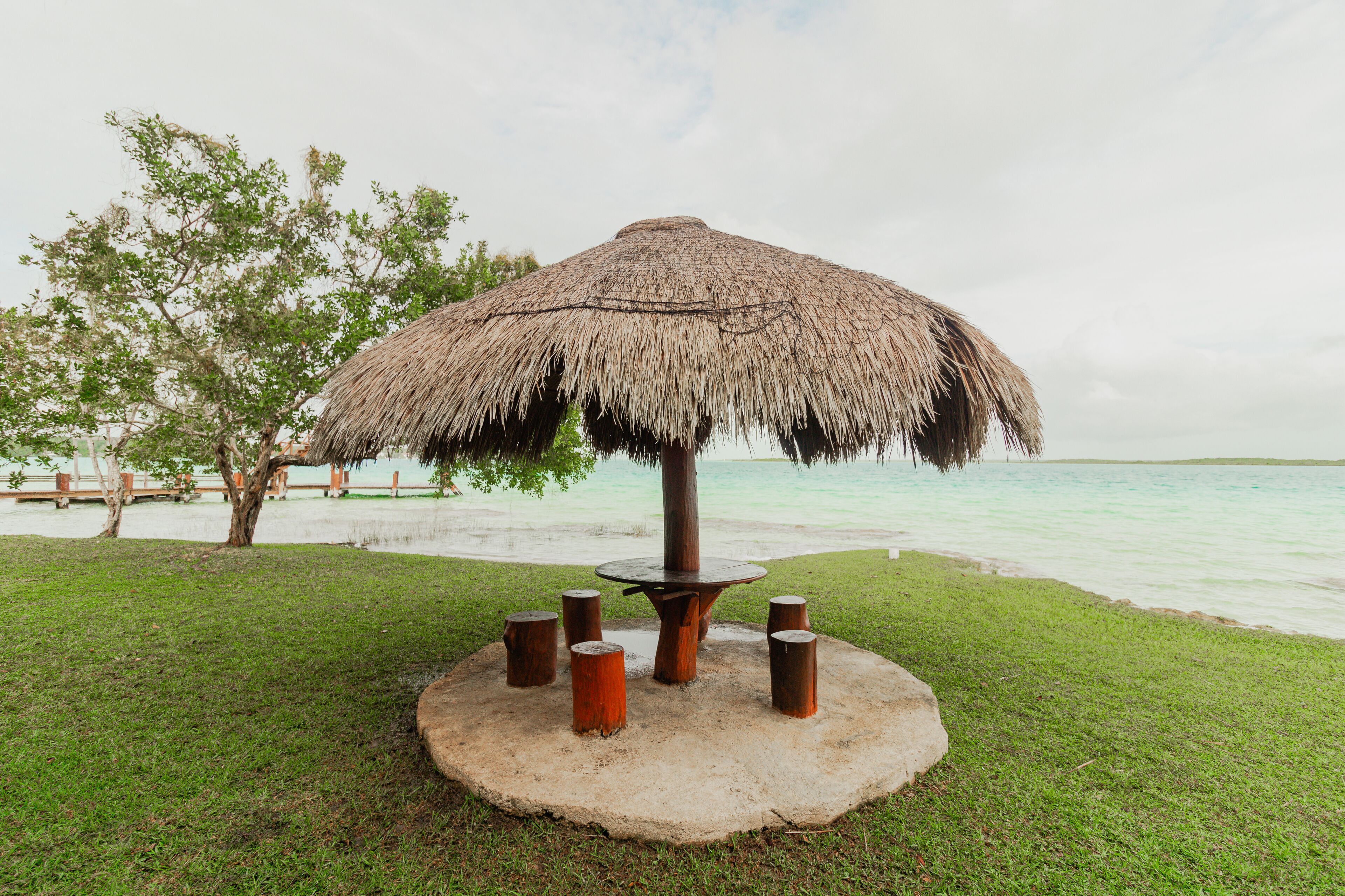 A thatched umbrella table with a circular base, surrounded by five wooden stools, set on a grassy area with a view of a calm body of water and trees in the background.