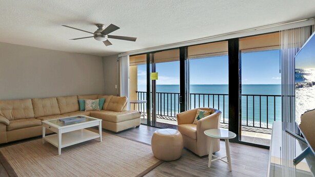 Ein geräumiges Wohnzimmer mit einem beige-farbenen Sofa, einem Couchtisch und einem runden Hocker. Es gibt einen Deckenventilator, große Fenster mit Meerblick und einen Balkon mit Geländer.