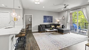 A modern kitchen with a white island and bar stools, a gray sofa with a coffee table, and a large window with curtains. - Chic 2BR Apt, Steps from N Main St, Balcony View
 (Greenville)