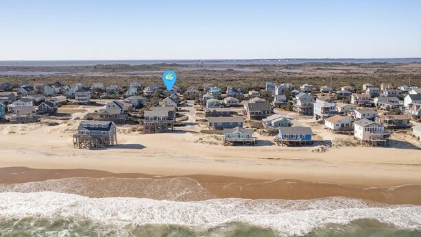 A coastal residential area with houses on stilts over the beach, a clear sky, and waves approaching the shore.