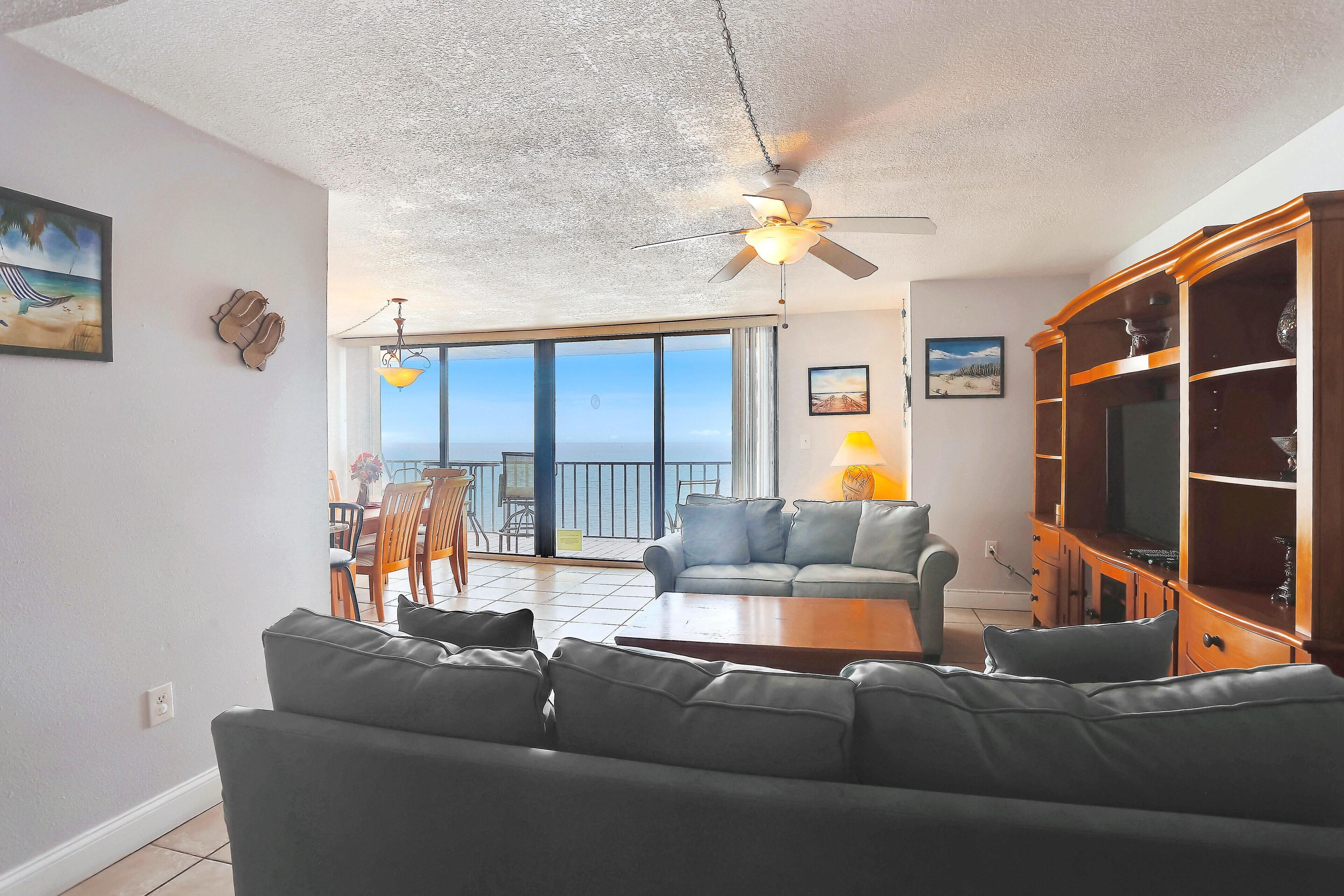 A living room with a sofa, a coffee table, a TV cabinet, and a ceiling fan. There is a balcony with a view of the ocean.