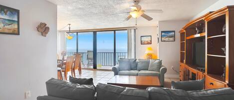 A living room with a sofa, a coffee table, a TV cabinet, and a ceiling fan. There is a balcony with a view of the ocean.