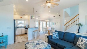 A spacious living area with a blue sofa, a patterned ottoman, a ceiling fan, and a kitchen in the background. - Pet-friendly oceanside home in Corolla w/ pool and hot tub! (Corolla)