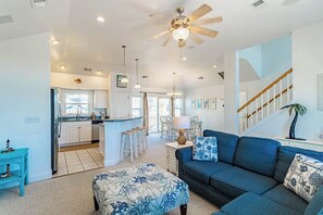 A spacious living area with a blue sofa, a patterned ottoman, a ceiling fan, and a kitchen in the background.