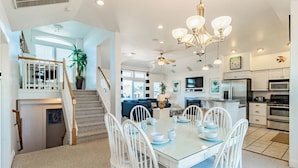 A spacious kitchen with modern appliances, a dining area with white chairs, and a staircase leading to a loft. - Pet-friendly oceanside home in Corolla w/ pool and hot tub! (Corolla)