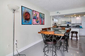 A modern kitchen with white cabinets, a central island, and a dining area with a glass table and metal chairs. - Trillium 5C - Premier (Madeira Beach)