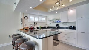 A modern kitchen with a central island, bar stools, white cabinetry, and a large window with blinds.