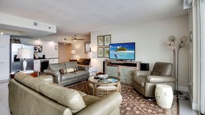 A modern living room with a sofa, armchair, and coffee table. There is a television mounted on the wall, a kitchen area in the background, and a decorative plant.