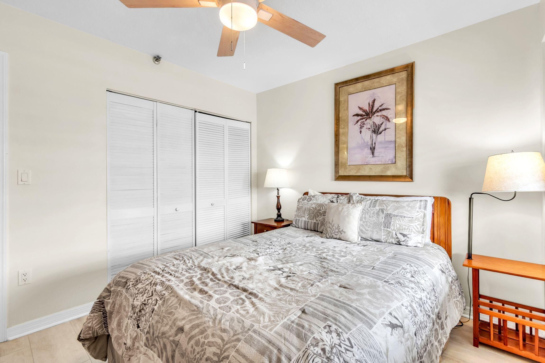 Quarto com cama, duas luminárias de cabeceira, ventilador de teto, quadro na parede e guarda-roupa com portas de correr.
