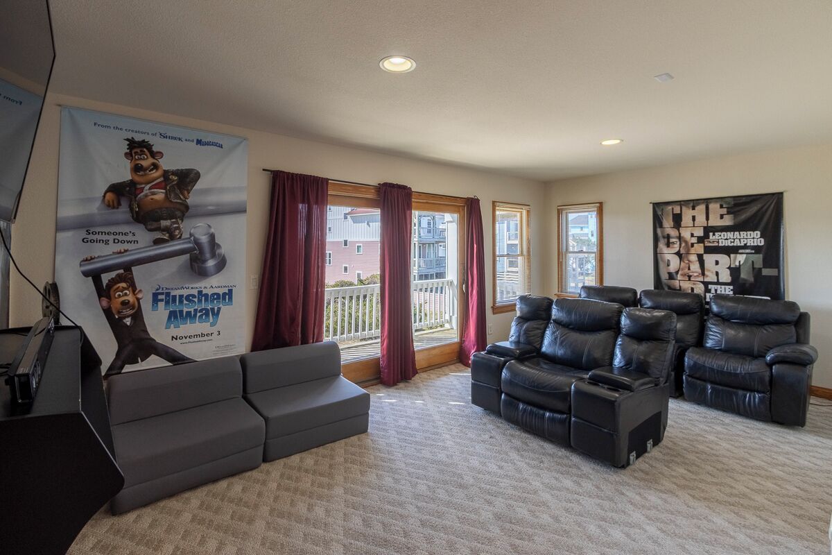A living room with a gray sectional sofa, black leather recliners, a large poster, and a balcony with red curtains.