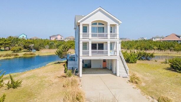 A two-story building with a balcony, a concrete pathway leading to the entrance, and a body of water in the background. - Spacious pet-friendly home on Hatteras Island w/ a private pool & hot tub! (Avon)