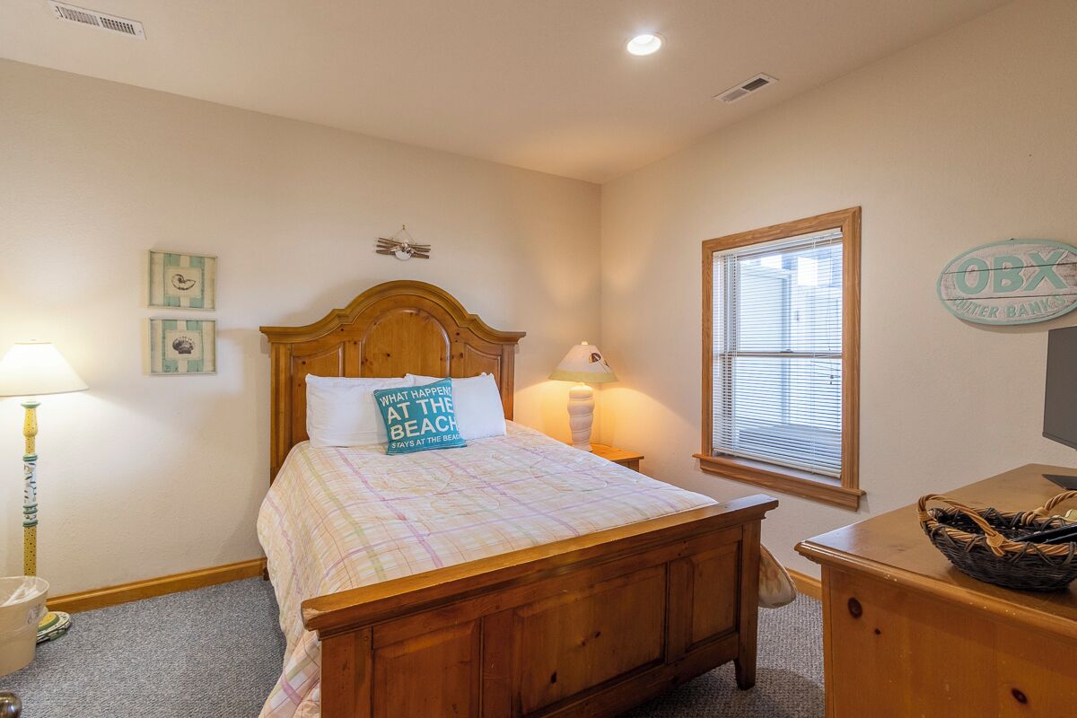 A bedroom with a wooden bed, a nightstand, a lamp, a window with blinds, and a wall sign.
