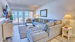 A spacious living room with a grey sectional sofa, a white side table with a lamp, a white cabinet, and a large window offering a view of the outdoors. - Villa Madeira 203 - Premier (Madeira Beach)