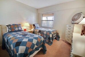 Une chambre avec deux lits, une commode, une lampe, une fenêtre avec des stores et un ventilateur sur pied.