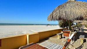 A beachside terrace with wooden lounge chairs, a thatched umbrella, and a view of the beach and ocean. - Villa Madeira 301 - Premier (Madeira Beach)