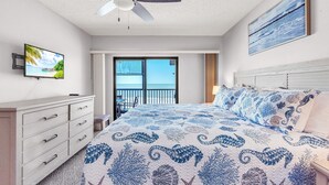 Ein Schlafzimmer mit Bett, Deckenventilator, Fernseher, Balkon mit Meerblick und einem Meeresbild.