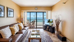 A living room with a brown leather sofa, a glass coffee table, a TV on a black stand, and a view of the ocean through a sliding glass door.
