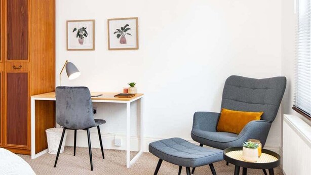 Ein Zimmer mit Holzschrank, Schreibtisch und Stuhl, grauem Sessel mit gelbem Kissen, rundem Tisch mit Pflanze und zwei gerahmten Bildern an der Wand.