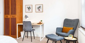 A room with a wooden wardrobe, a desk with a chair, a grey armchair with a yellow pillow, a small round table with a plant, and two framed pictures on the wall.