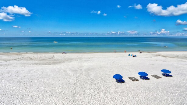 A beach with white sand and a clear blue sky. There are several blue umbrellas and lounge chairs. - Villa Madeira 406 (Madeira Beach)