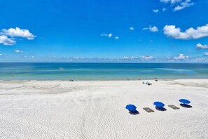 Ein Strand mit weißem Sand und blauem Himmel. Mehrere blaue Sonnenschirme und Liegestühle sind zu sehen.