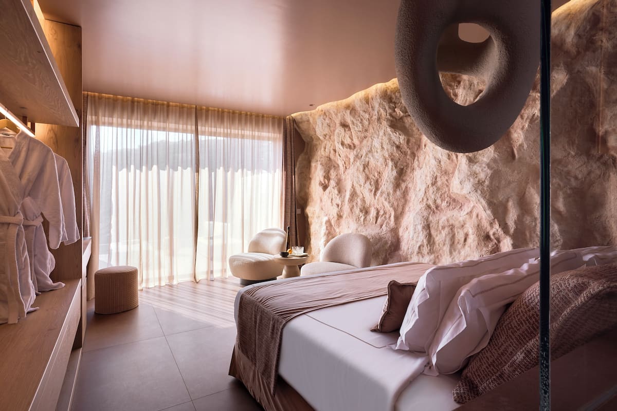 A modern bedroom with a large bed, a round hanging light fixture, a seating area with a small table, and a large window with sheer curtains.