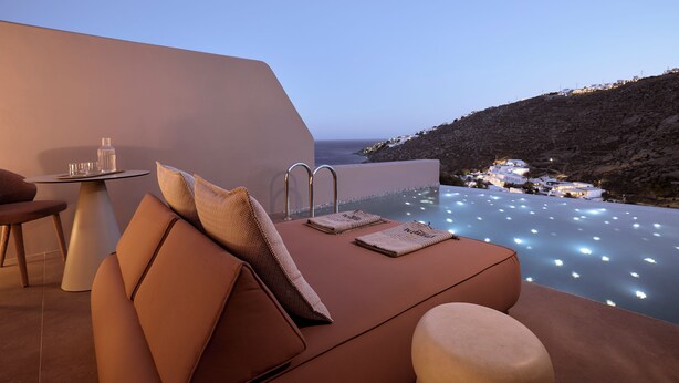 Un área de estar exterior moderna con piscina, una mesa con vasos y una botella, y vistas de la costa al atardecer.