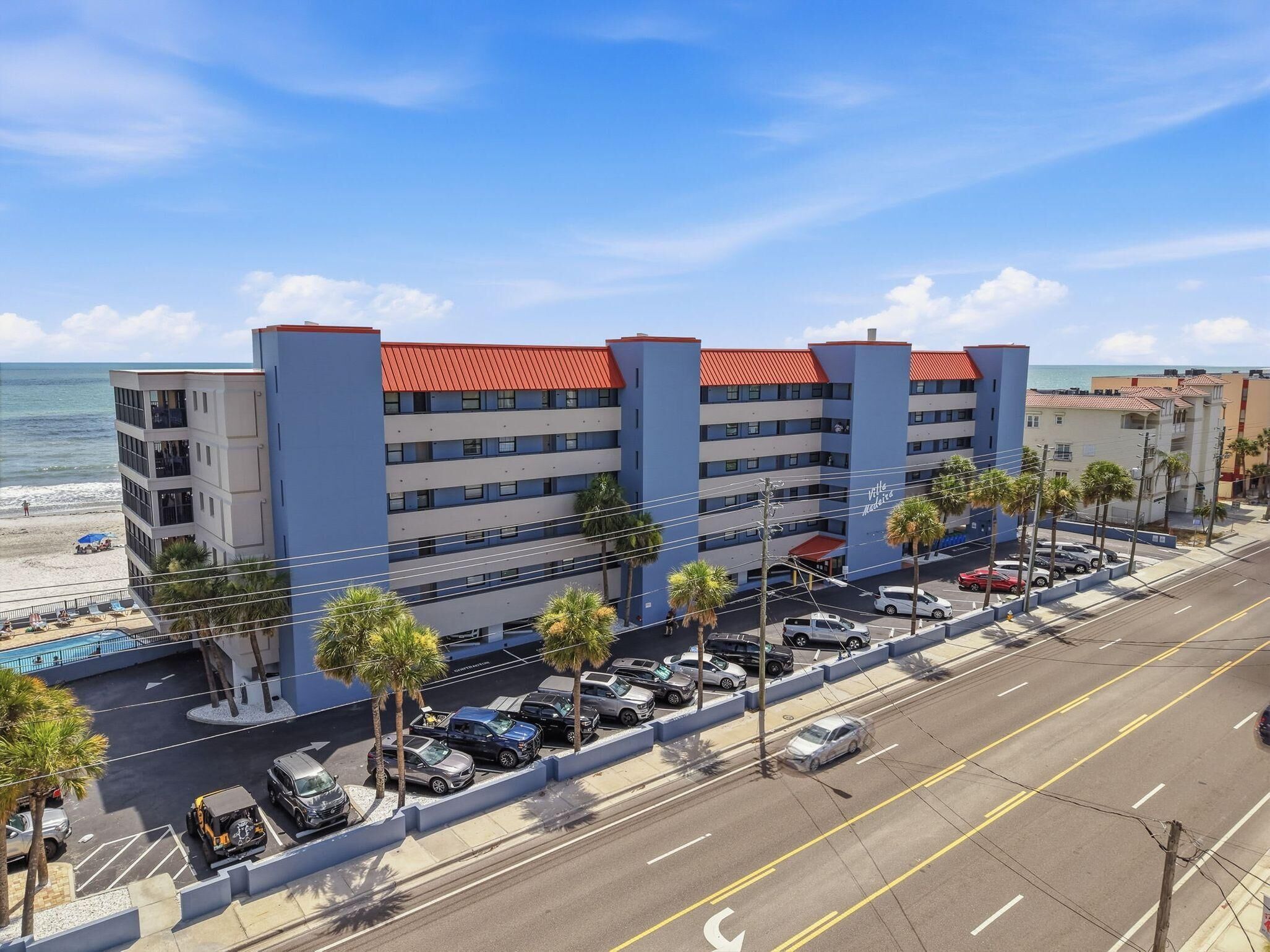 A multi-story hotel with a red roof, located near a beach with visible palm trees and parked cars.