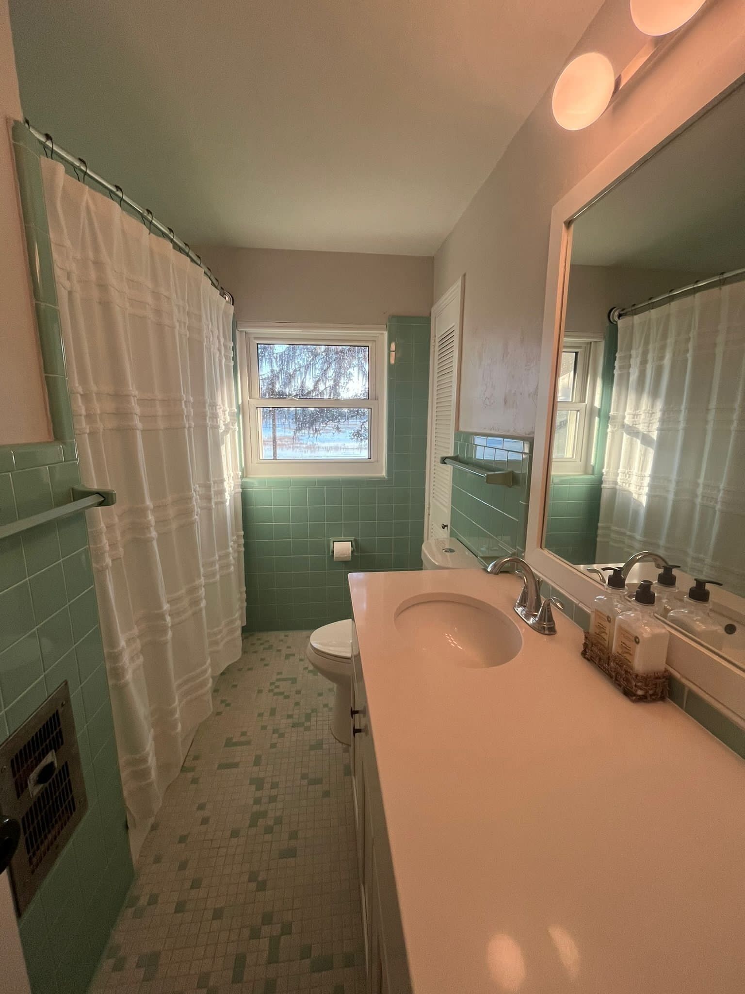 Une salle de bain avec des toilettes, un lavabo et un grand miroir. Il y a une fenêtre donnant sur des arbres et un rideau de douche.
