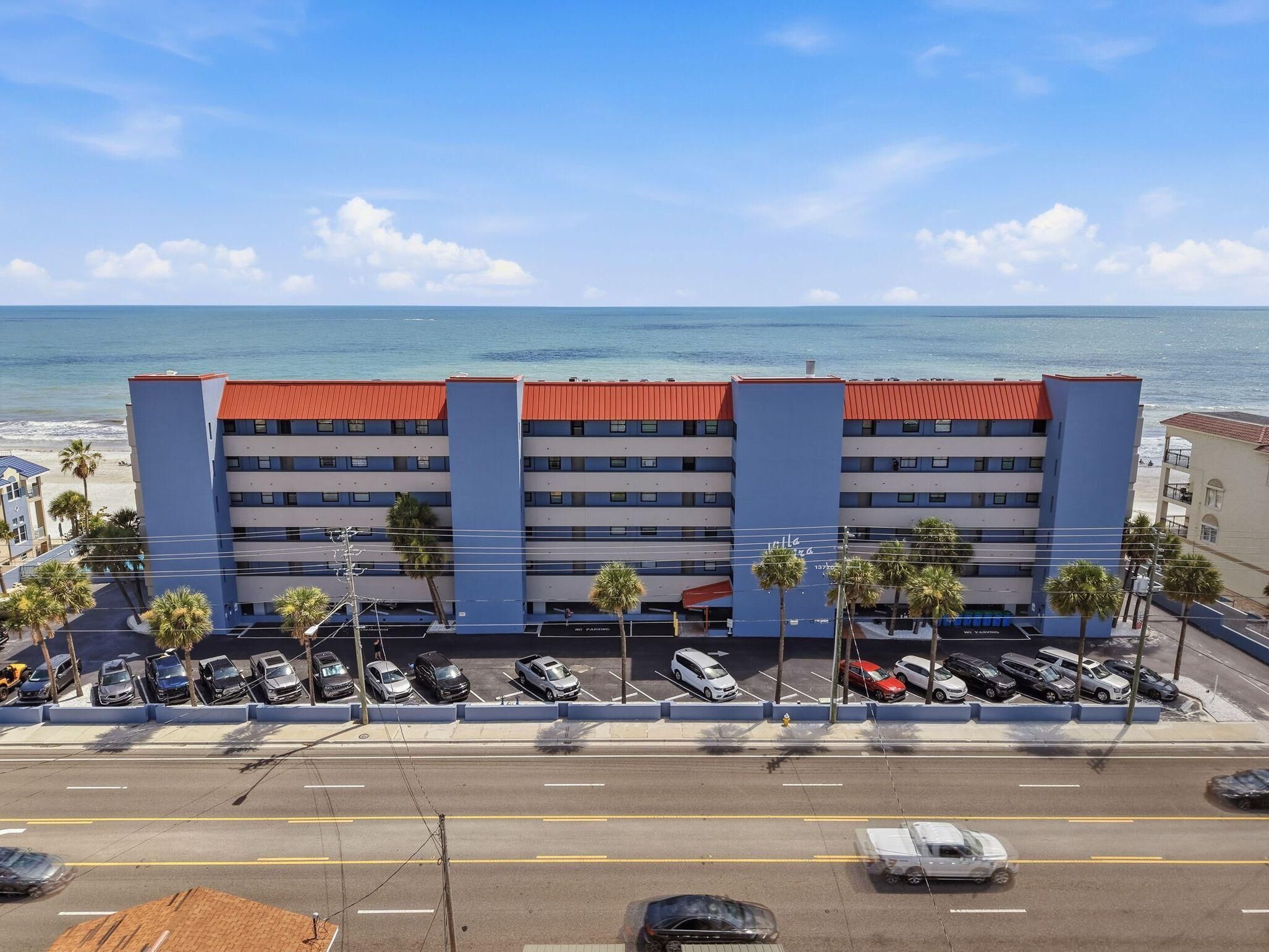 A multi-story hotel with a red roof, parking lot, and a view of the ocean.