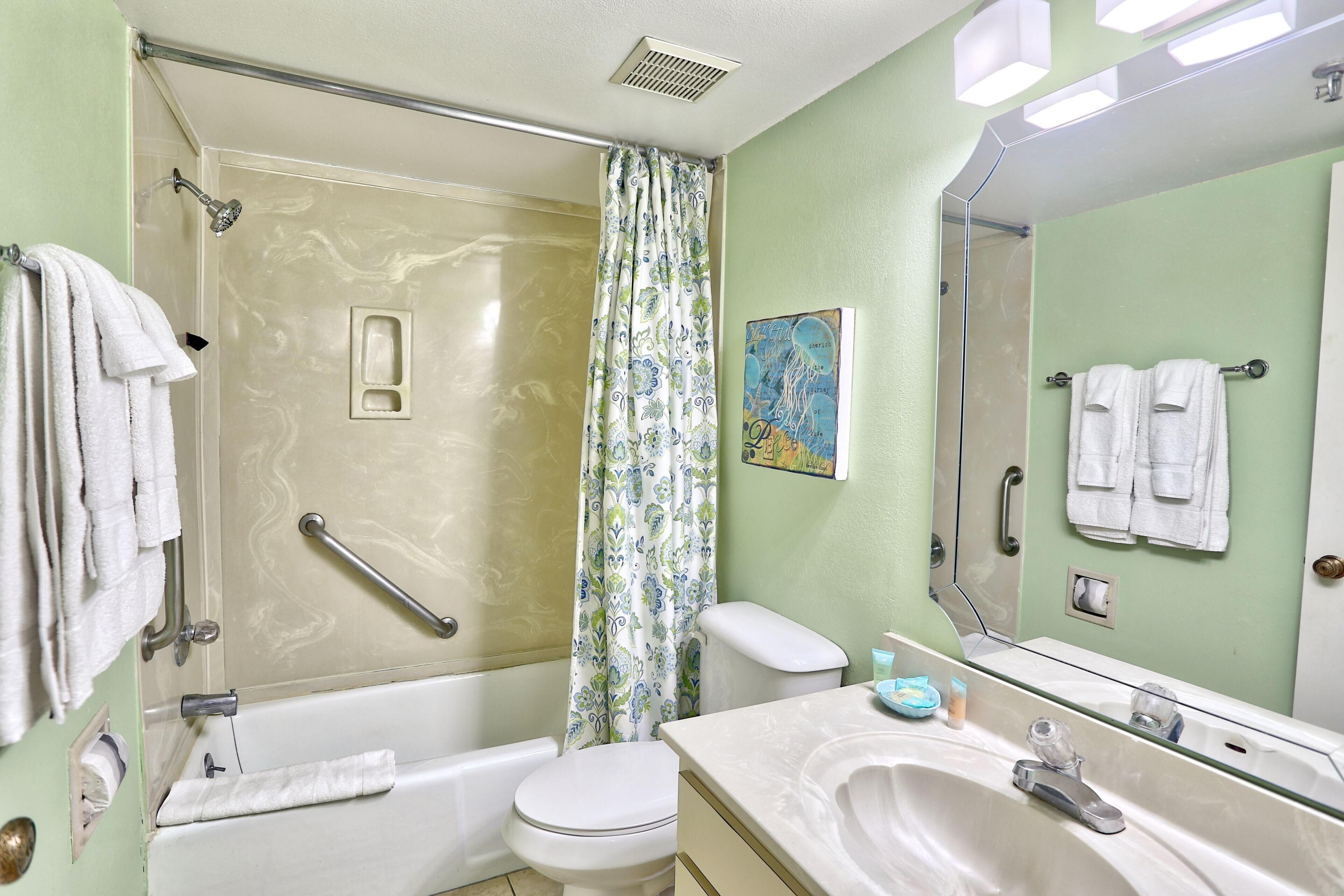 A bathroom with a shower, toilet, and sink, equipped with a handrail and a mirror.