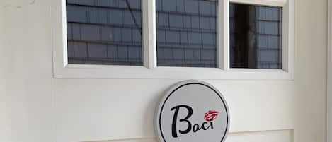 Une porte blanche avec une fenêtre au-dessus et un panneau circulaire portant le mot « Baci » et un motif de lèvres rouges.