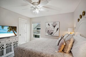 A bedroom with a bed, a ceiling fan, a TV, and a window with a view of a beach.