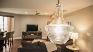 Een moderne woonkamer met een glazen hanglamp, een grijze bank, een eethoek met stoelen en een televisie op de achtergrond.