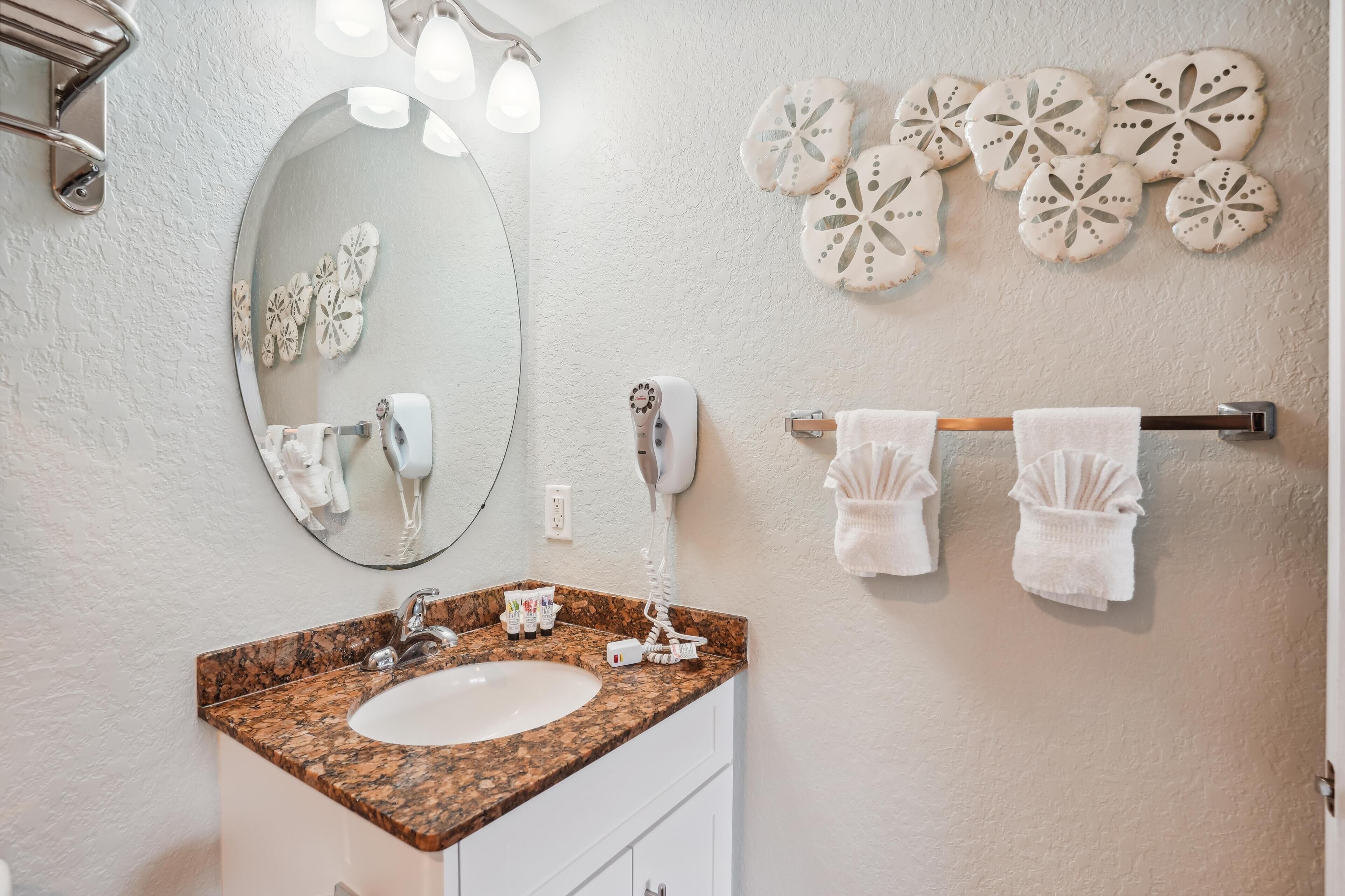 Une salle de bain dotée d’un lavabo avec un plan de toilette en granit, d’un miroir rond, d’un sèche-cheveux et d’œuvres d’art décoratives murales.