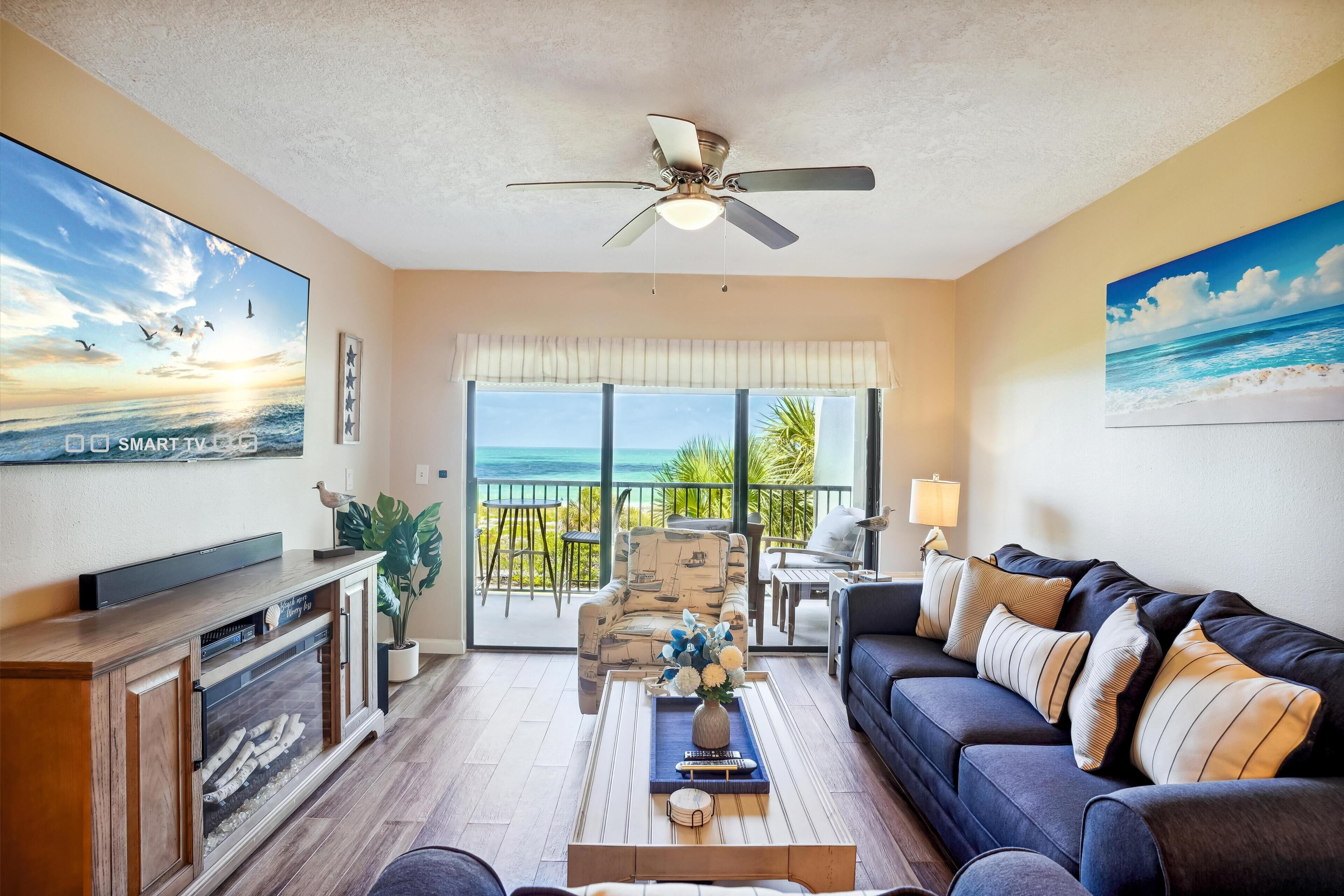 A living room with a flat-screen TV, a ceiling fan, a sofa, a coffee table, and a view of the ocean through large windows.