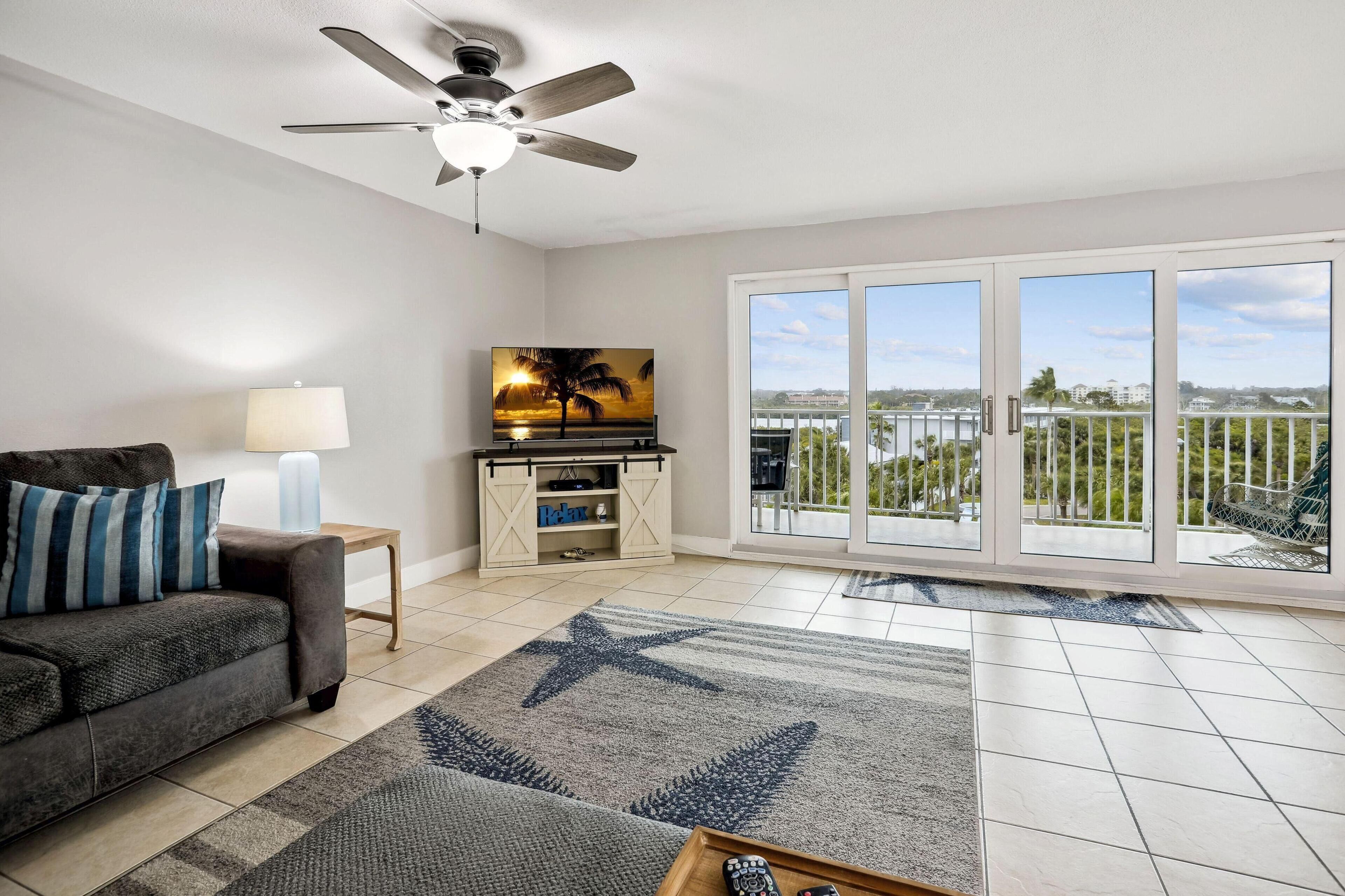 A living room with a sofa, a TV, a ceiling fan, and a large window with a view of the city.