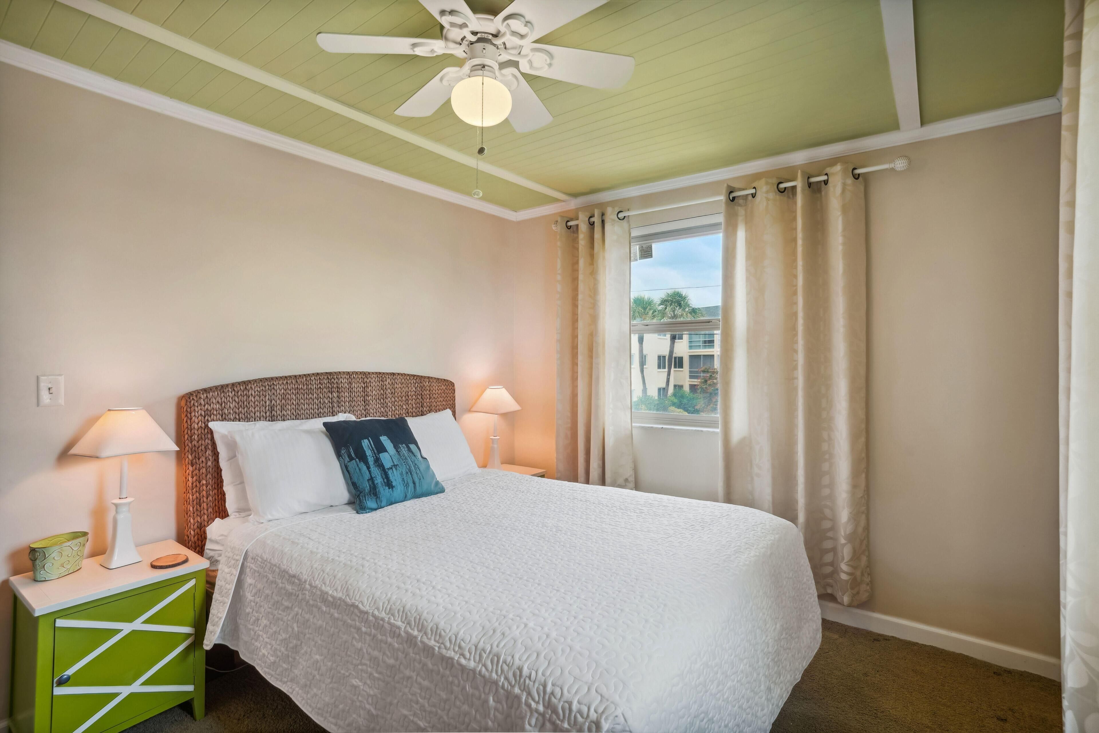 A bedroom with a bed, two bedside lamps, a green nightstand, a ceiling fan, and a window with curtains.