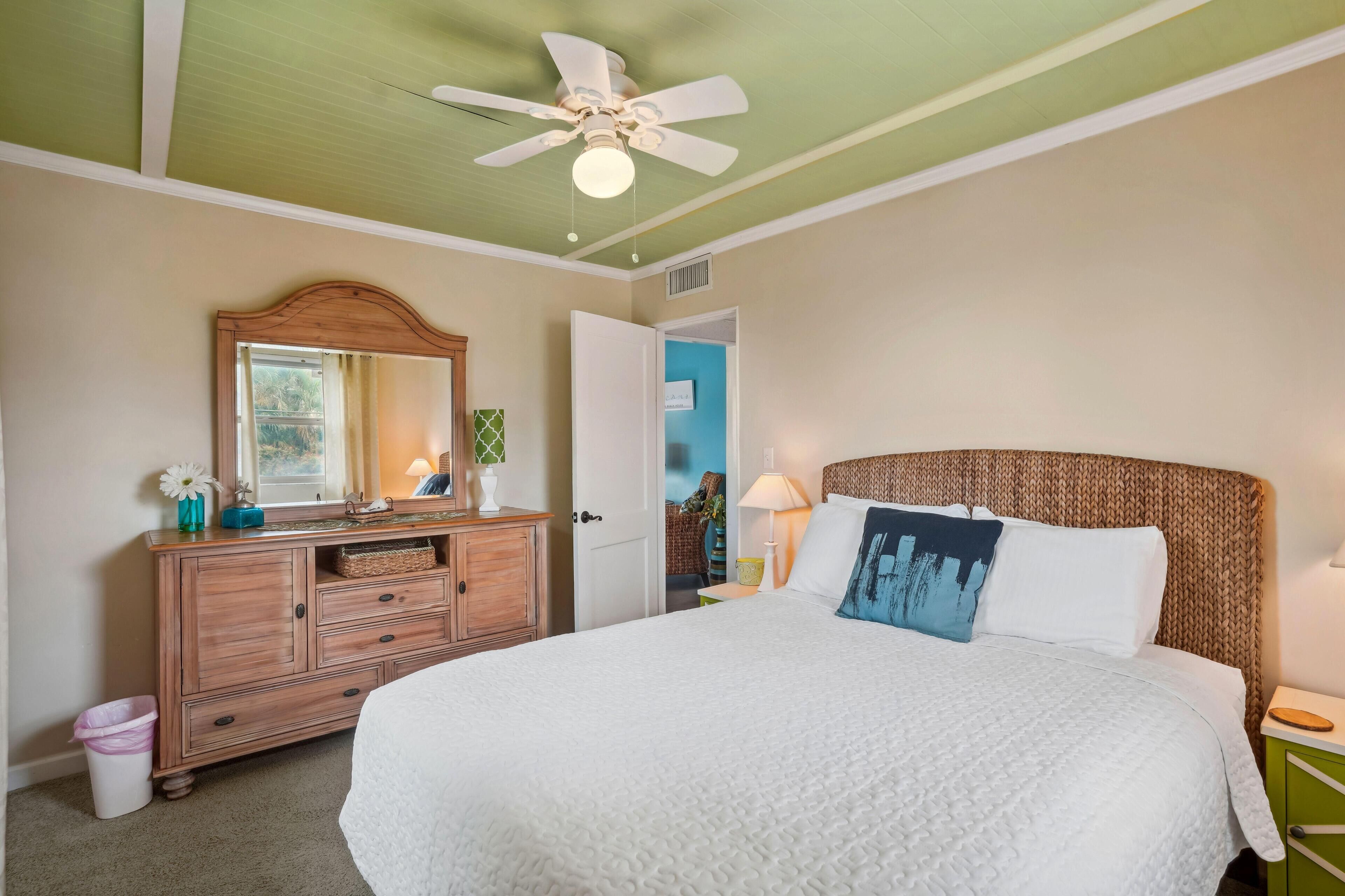 Une chambre à coucher avec un grand lit, une commode en bois, un ventilateur de plafond et une porte donnant sur une autre pièce.