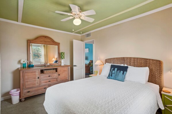A bedroom with a large bed, a wooden dresser, a ceiling fan, and a door leading to another room. - Friendly Native 507 (St. Pete Beach)