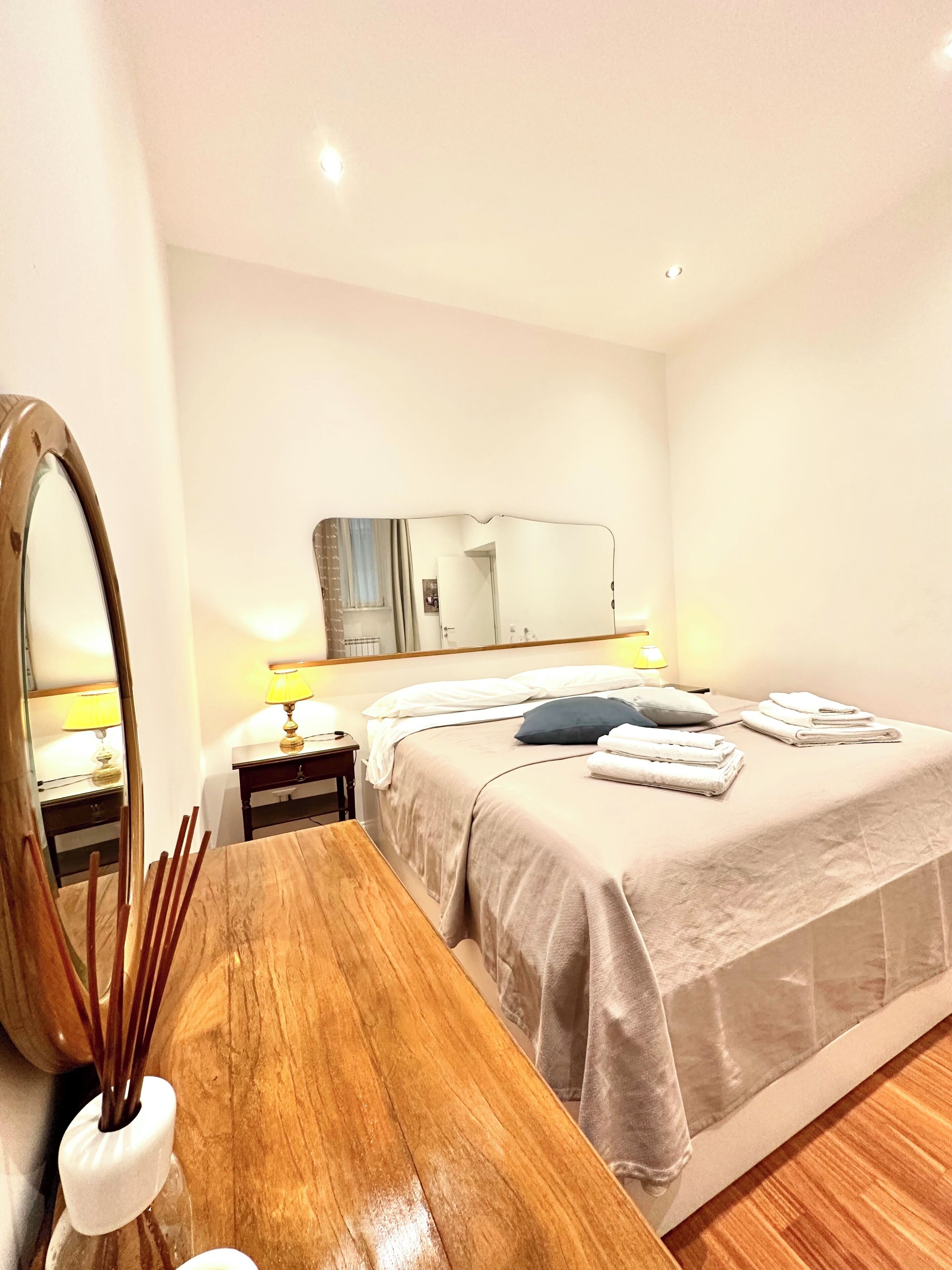 Een hotelkamer met een houten bureau, een grote spiegel, een bed met een beige dekbedovertrek en een nachtkastje met een lamp.