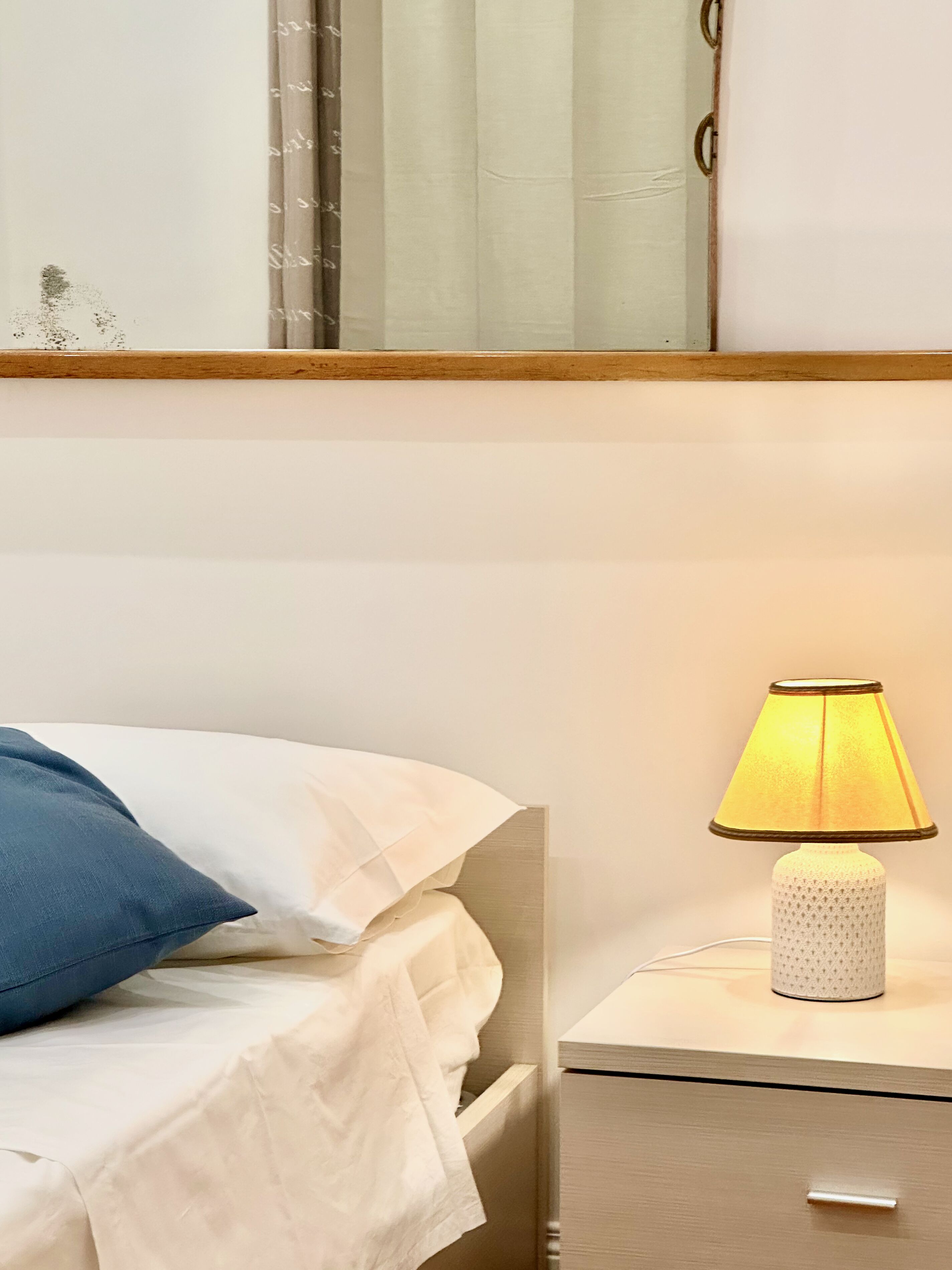A neatly made bed with a blue pillow and a white blanket, a bedside lamp, and a small drawer unit.