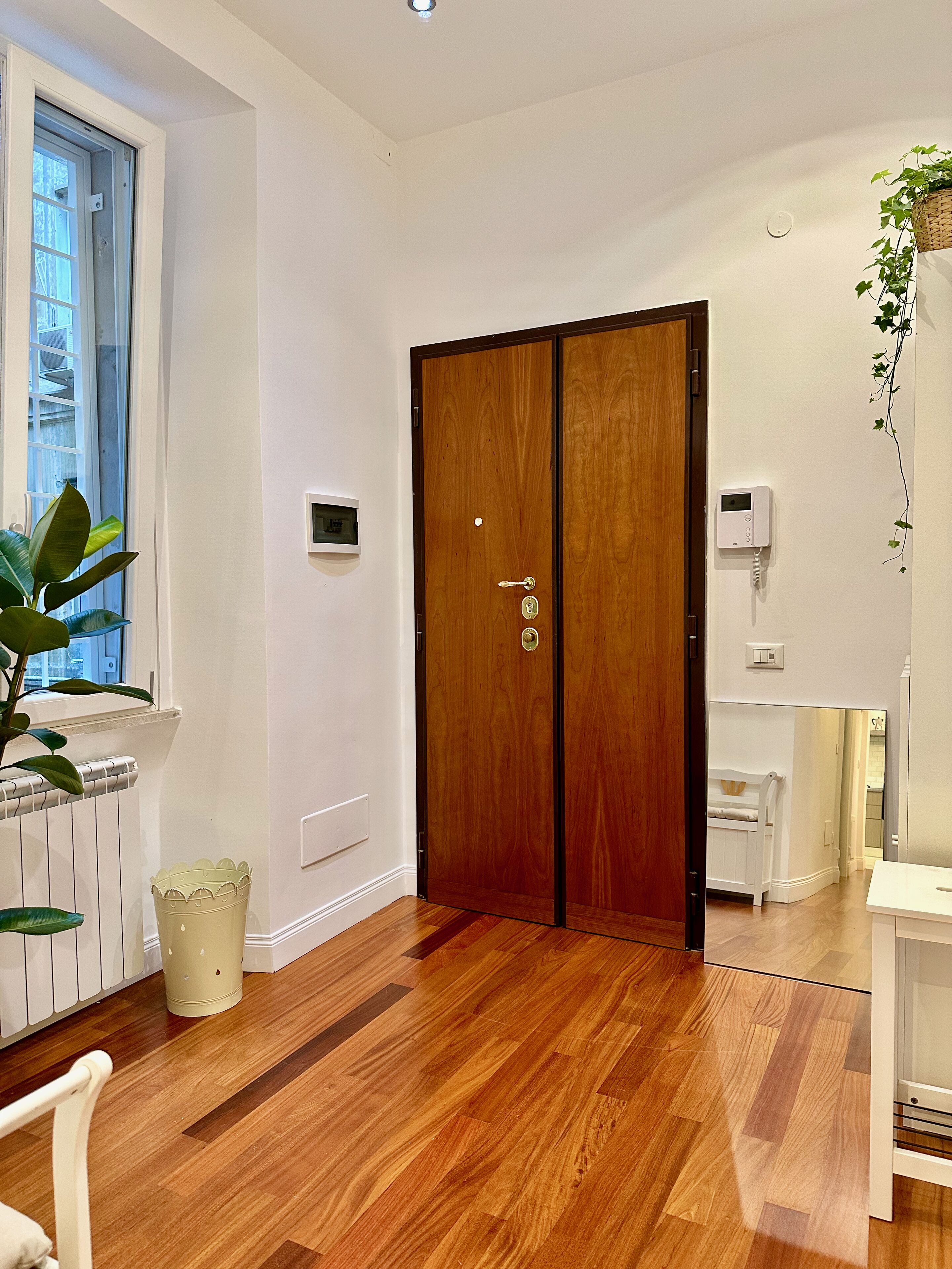 A hallway with wooden flooring, a double wooden door, a radiator, a potted plant, and a white wall with a thermostat.