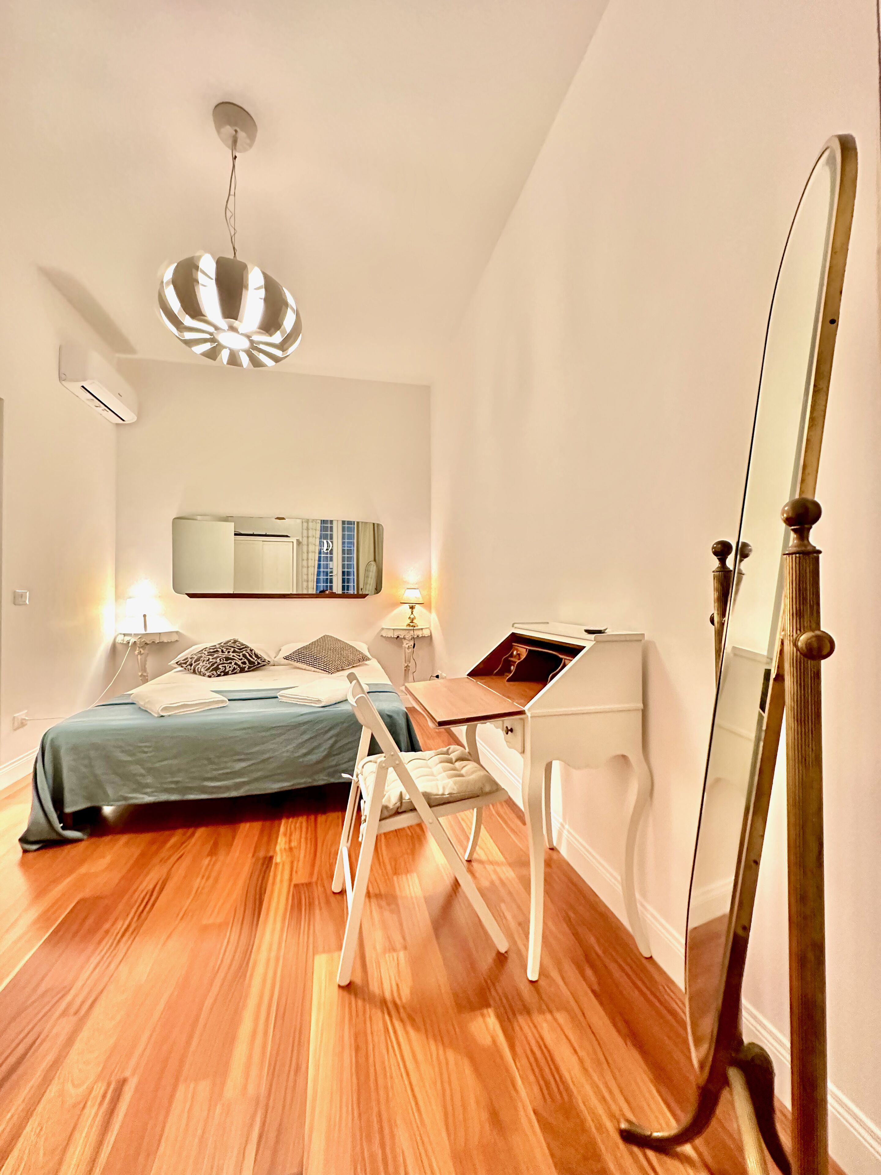 A modern bedroom with a bed, bedside tables, a desk, a mirror, and a hanging light fixture.
