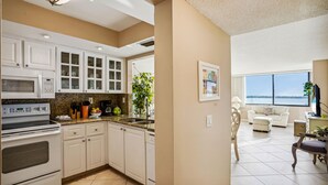 Una cocina con armarios blancos, una estufa y un lavabo. Hay una zona de comedor con un sofá blanco y vistas al exterior.