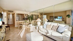 Amplia sala de estar con un sofá blanco, una mesa de comedor y sillas. Se observa una cocina al fondo, y un espejo grande refleja la habitación.