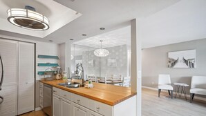 A modern kitchen with a wooden countertop, white cabinets, and a large island. There is a dining area with white chairs and a small table. - Land's End 1-306 Bay Front- Premier (Treasure Island)