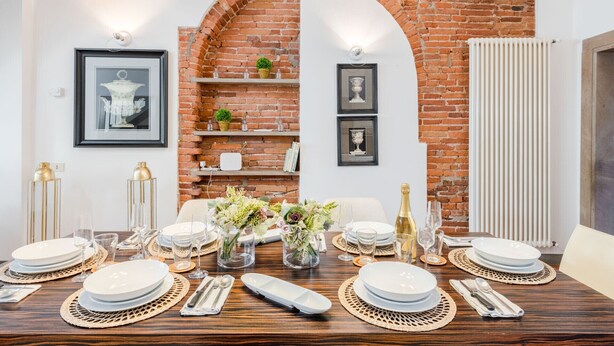A dining area with a brick wall, white walls, and a white radiator. There are framed pictures on the wall, a table set with plates, glasses, and cutlery. - Modern Panoramic Penthouse with Elevator inside the Walls of Lucca (Lucca)