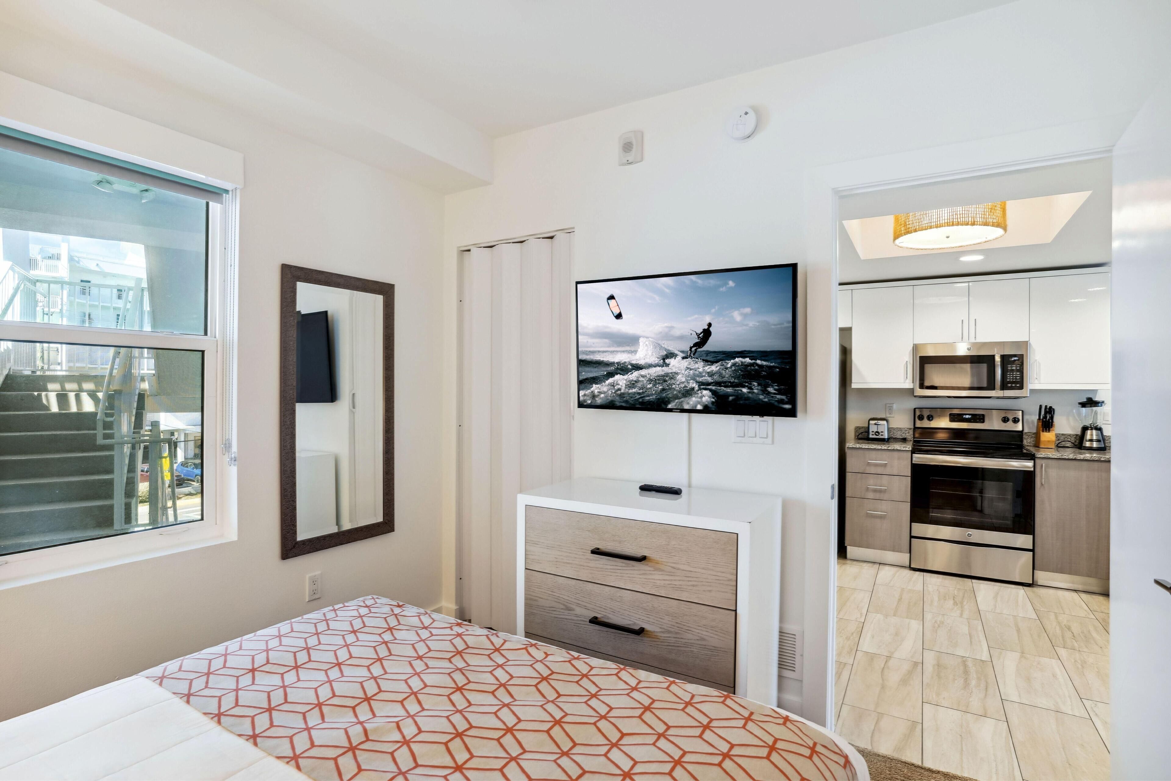 A modern hotel room with a bed, a TV, a kitchenette, and a view of the outside.