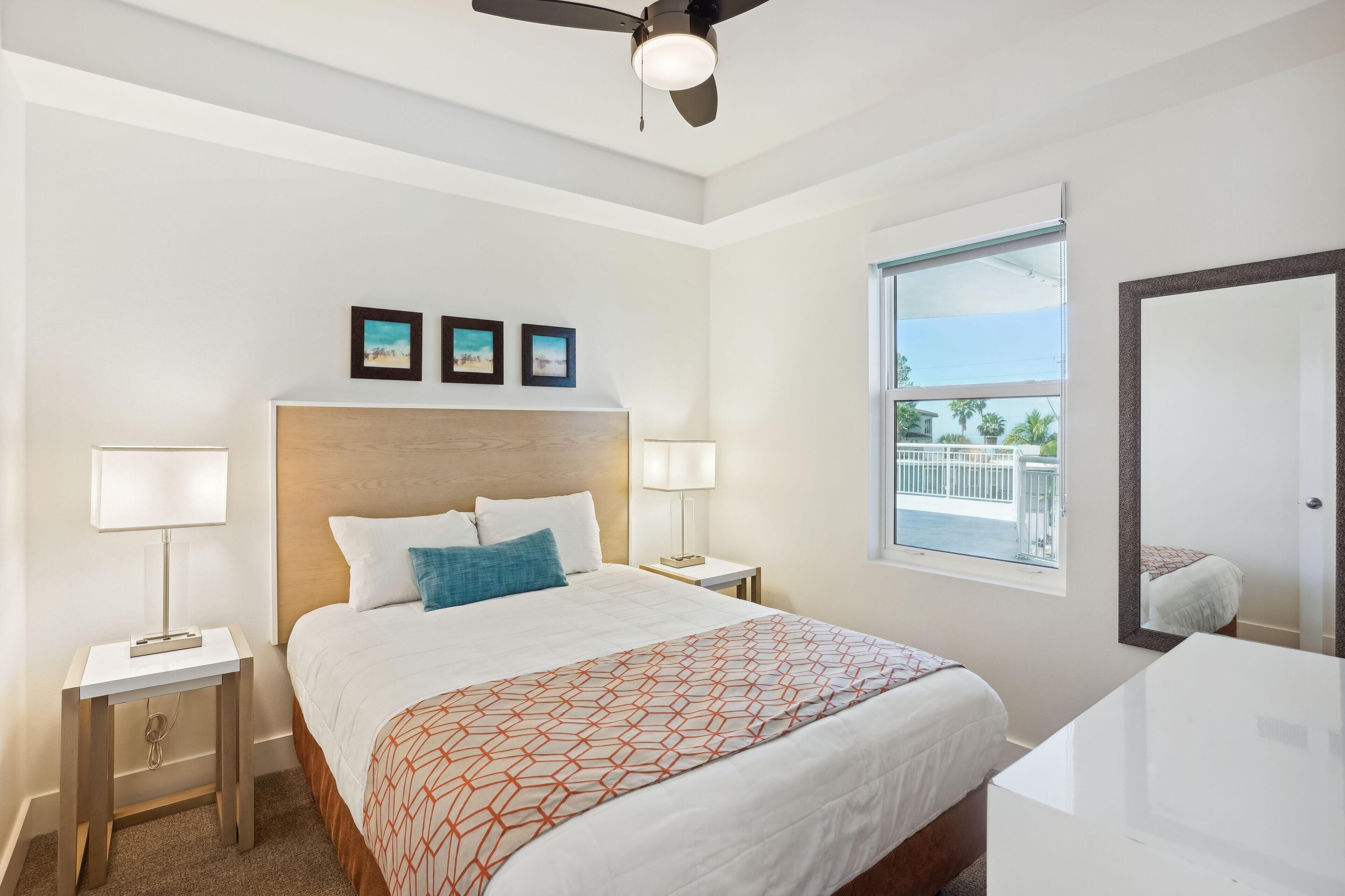 Habitación de hotel con cama, lámparas de noche, ventilador de techo, ventana con vista al balcón y baño con espejo y lavamanos.